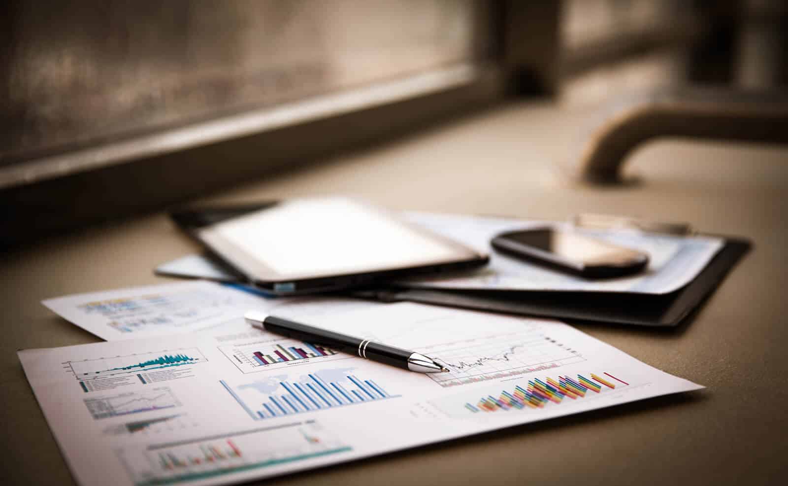 Pöydällä on taloudellisia raportteja, joissa näkyy kaavioita ja analytiikkaa, sekä mustekynä, tabletti ja älypuhelin. Kuva symboloi yritysrahoituksen suunnittelua, investointien hallintaa ja liiketoiminnan kehittämistä. Se viittaa muun muassa yrityslainaan, laskurahoitukseen ja luotonvalvontaan, jotka tukevat yrityksen taloudellista kasvua.